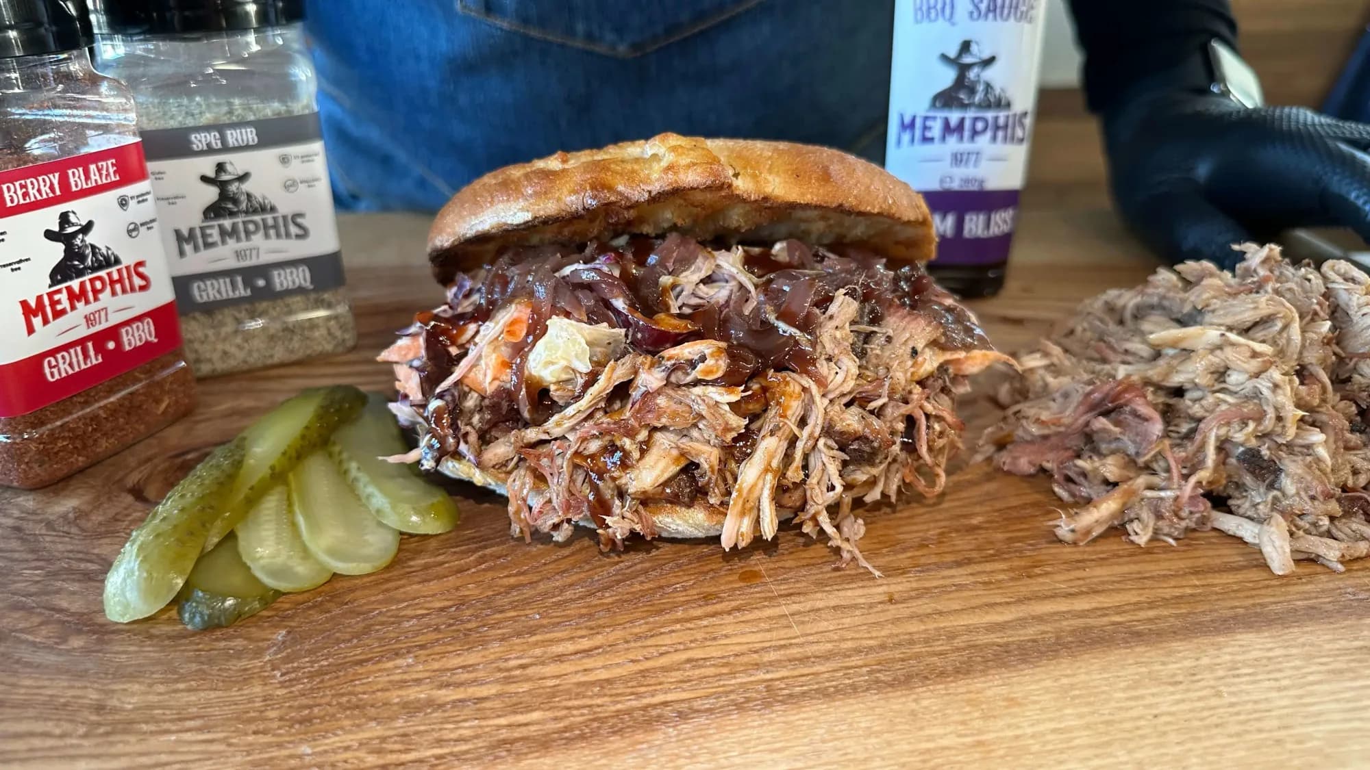 Sandwich Pulled Pork gatit in gratar ceramic Kamado pe un tocator masiv din lemn cu condimente BBQ Memphis si cu sos BBQ Memp
