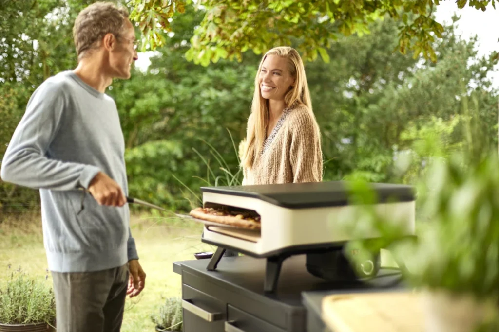 Cuptor pizza in aer liber pe un blat intr-o bucatarie exterioara cu pizza delicioasa scoasa cu o paleta de pizza si inconjurata de prieteni si familie